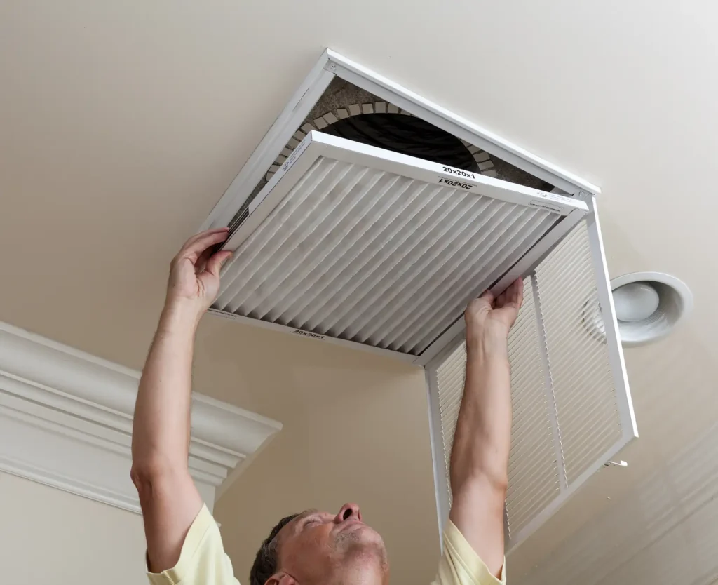 Person lifting a pleated HVAC air filter into an open ceiling return vent during indoor air maintenance.